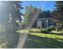 Ein Garten mit frisch gemähtem Rasen, umgeben von Bäumen und Büschen, im Vordergrund steht eine Sonnenliege. Im Hintergrund ist ein kleines, modernes Gartenhaus mit offener Glastür und Flachdach zu sehen.