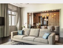 Wohnzimmer einer modernen Wohnung mit grauem Sofa im Vordergrund, dekoriert mit Kissen. Im Hintergrund ein Essbereich mit hölzernen Wänden, einem Esstisch und gepolsterten Stühlen. Ein auffälliger Kronleuchter mit goldenen Kugeln hängt über dem Tisch.