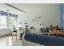 Helles Jugendzimmer mit Holzfußboden, einem Schreibtisch vor einem großen Fenster, das Tageslicht hereinlässt, und einem Bett mit Kissen und Stofftieren auf der rechten Seite. An der Wand befinden sich ein Pinnbrett, Regale und verschiedene Dekorationen.