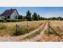 Ein einzelnes weißes Einfamilienhaus mit dunklem Dach steht am Rand einer ländlichen Wiese, durch die ein unbefestigter Feldweg führt. Rechts und links des Weges sind hohe Gräser, eingezäunt von einfachen Metallpfosten, im Hintergrund sind Bäume sichtbar.
