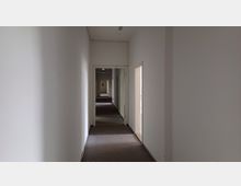 Ein schmaler, langer Flur in einem Gebäude mit weiß gestrichenen Wänden und Teppichboden. Mehrere offene Türen führen in angrenzende Räume, und der Flur erstreckt sich in gerader Linie nach hinten.