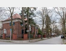 Straßenszene mit einem roten Backsteinhaus mit blau gestrichenen Fensterläden und einem roten Ziegeldach auf der linken Seite, entlang einer gepflasterten Straße. Mehrere Bäume säumen den Gehweg, und Straßenschilder mit den Namen „Gelbertstraße“ und „Flüthestraße“ sind sichtbar.