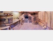 Ein traditioneller Backraum mit einem gemauerten Backofen in der Mitte, umgeben von Holzwänden. Links stehen ein Tisch mit Backutensilien und Regalen, rechts Stapel aus Holz und gestapelte Stühle; der Boden besteht aus Backsteinen.