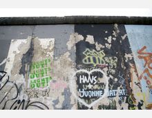 Abschnitt einer alten Betonwand mit bröckelnder Farbe und diversen Graffiti, darunter Schriftzüge wie "Hans + Yvonne" in einem weißen Herz sowie "Lights Out!" in grün. Die Oberfläche zeigt Abnutzungsspuren und farblich verblasste Bereiche.