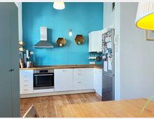 Moderne Küche mit Arbeitsplatte aus Holz, weißen Schränken und eingebautem Herd. Die Rückwand ist türkis gestrichen und mit zwei hexagonalen Holzwandregalen dekoriert, rechts steht ein Kühlschrank mit Magneten und Notizen.