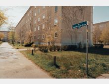 Wohngebiet mit einem mehrstöckigen Plattenbau auf der linken Seite, umgeben von vereinzelten Bäumen und Grünflächen. Im Vordergrund steht ein Straßenschild mit der Aufschrift „Zur Friedrich-Engels-Straße“.