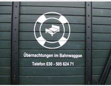 Dunkelgrüne Holzwand eines historischen Bahnwagens mit weißem Logo eines Betts in einem Rettungsring und der Aufschrift „Übernachtungen im Bahnwaggon“ sowie einer Telefonnummer (030 - 505 624 71). Metalldetails und Holzverkleidung sind sichtbar.