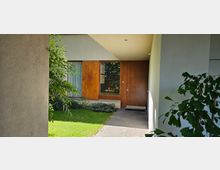 Eingangsbereich eines modernen Hauses mit einer Holzhaustür und einem großen Fenster mit Vorhang. Der Bereich ist von Grünpflanzen umgeben, mit einem kleinen Rasen und Pflastersteinen vor der Tür.