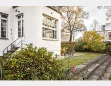 Außenansicht eines weißen Hauses mit großen Sprossenfenstern und einer kleinen Treppe mit Metallgeländer, die zu einer Seitentür führt. Im Vorgarten befinden sich Büsche, blühende Pflanzen, ein gepflegter Rasen und eine Sitzgruppe neben einem Baum in herbstlicher Umgebung.