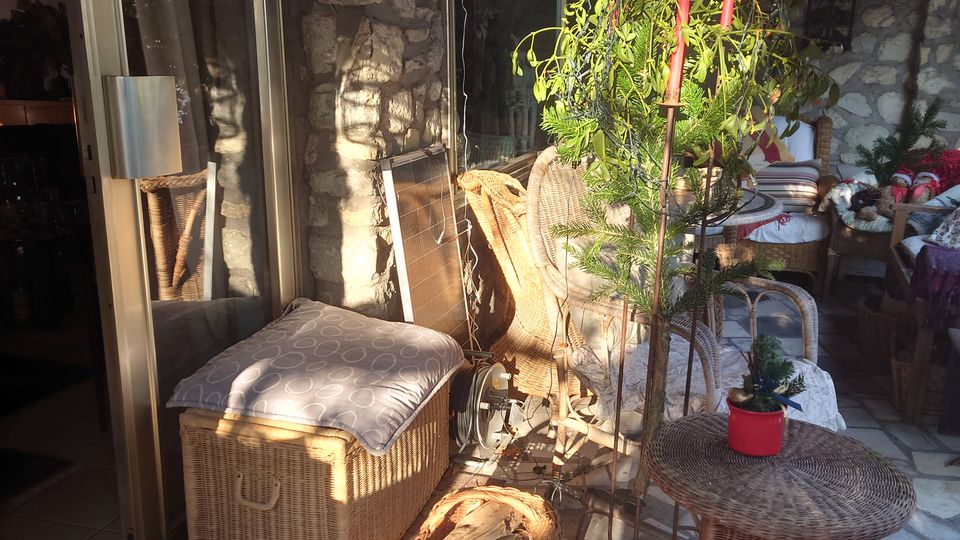 Steinerne Terrasse mit Glas-Schiebetür, rustikal eingerichteten Korbmöbeln, Pflanzen in Töpfen und einem gefliesten Boden. Die Wand besteht aus grobem Naturstein, und die Szenerie wird von Sonnenlicht erhellt.