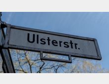 Straßenschild mit der Aufschrift "Ulsterstr." in schwarzer Schrift auf weißem Hintergrund, umgeben von einem Metallrahmen; im Hintergrund sind Äste von Bäumen und ein blauer Himmel zu sehen.