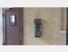 Holz-Tür mit abblätterndem braunem Lack und kleinem Fenster in einer gelb gestrichenen Wand; daneben ein verwittertes, grünlich-blaues Kästchen mit Rostspuren und eine Steckdosenleiste.