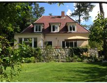 Zweistöckiges Einfamilienhaus mit gelber Fassade, roten Ziegeldach und mehreren Dachgauben, umgeben von einem gepflegten Garten mit Rasen, Bäumen und einer Terrasse. Auf der Terrasse steht ein großer Sonnenschirm, und eine niedrige Steinmauer mit Pflanzen begrünt den Bereich.