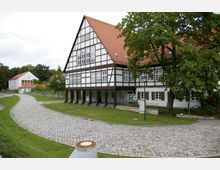 Fachwerkhaus mit weißen Wänden und dunklen Holzbalken, rotem Satteldach und zahlreichen Fenstern, umgeben von einer grünen Wiese und gepflasterten Wegen. Im Hintergrund stehen weitere Häuser und Bäume.