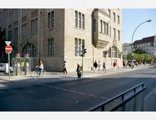 Straßenszene mit einem großen historischen Gebäude aus hellem Stein und hohen Fenstern auf einer Ecke. Menschen gehen oder fahren mit dem Fahrrad auf dem Gehweg, daneben befinden sich Telefonzellen und eine Ampel.