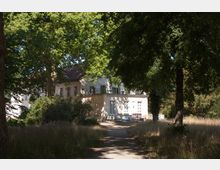 Helles, zweistöckiges Gebäude mit weißen Wänden und einem rötlichen Dach, umgeben von hohen Bäumen und einem naturbelassenen Garten. Ein schmaler Weg führt durch die Wiese auf das Gebäude zu.