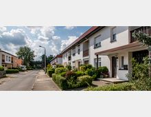 Reihenhäuser in einer ruhigen Wohnstraße mit Vorgärten, Hecken und Gehweg, umgeben von vereinzelt stehenden Bäumen und bewölktem Himmel.