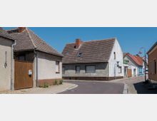 Eine kleine Straßenecke in einem Dorf mit einfachen, verputzten Häusern und Satteldächern. Der Straßenbelag wechselt von Asphalt zu Kopfsteinpflaster, und im Hintergrund ist eine laternenartige Straßenlampe zu sehen.