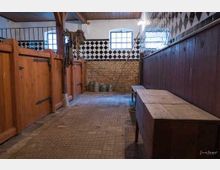 Innenansicht eines historischen Stallgebäudes mit Holztüren auf der linken Seite, gefliesten Wänden mit schwarz-weißem Rautenmuster, zwei Fenstern mit Gitterstäben und einem Holztisch auf der rechten Seite. Der Boden besteht aus einem gemusterten Steinbelag, und im Hintergrund sind Eimer erkennbar.