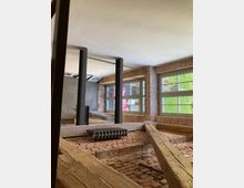 Innenansicht eines Gebäudes mit rustikaler Backsteinmauer und sichtbaren Holzbalken. Große Fenster lassen viel Tageslicht herein, während schwarze Metallträger und eine alte Heizvorrichtung den industriellen Stil betonen.