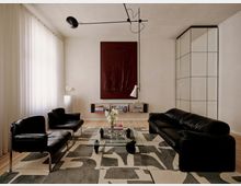 Modernes Wohnzimmer mit weißen Wänden, bodentiefen Vorhängen und hellen Holzdielen, eingerichtet mit schwarzen Leder-Sofas und Sesseln um einen Glas-Couchtisch auf einem grafisch gemusterten Teppich. An der Rückwand hängen ein großes dunkelrotes Kunstbild über einem niedrigen Regal, rechts steht ein hoher weißer Einbauschrank; eine minimalistische schwarze Deckenleuchte spannt sich über den Raum.