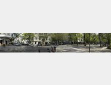 Eine städtische Straßenszene mit Altbauwohnungen, Cafés mit Außenbestuhlung und geparkten Autos entlang der Straße. Der Platz ist von Bäumen gesäumt und bietet offene Gehwege mit vereinzelten Laternen.