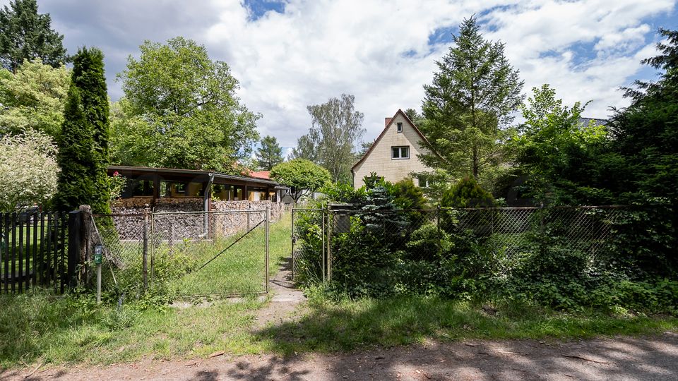 Blick auf ein eingezäuntes Wohngrundstück mit Metalltor und schmalem Weg, umgeben von dichtem Grün und hohen Bäumen. Im Hintergrund stehen ein kleines Haus mit steilem Giebeldach sowie ein niedriger Anbau mit Natursteinmauer und Flachdach.