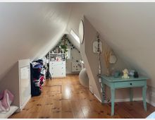 Schmales Dachgeschosszimmer mit schrägen weißen Wänden und Holzdielenboden, mit einem Oberlichtfenster und Möbeln entlang der Seiten. Rechts steht ein kleiner türkisfarbener Tisch mit Dekoration, links Regale und eine Kommode; in der Mitte führt ein freier Gang zum hinteren Bereich.