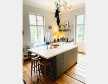 Moderne Wohnküche mit einer zentralen Kücheninsel mit weißer Marmorarbeitsplatte und Holzbarhockern, umgeben von hellen Wänden, großen Fenstern mit Tageslicht und Holzboden. An der Wand sind Regale mit Geschirr und Dekoration, eine Hängelampe sowie ein großer schwarzer Stern als Dekoration sichtbar.