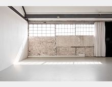 Ein minimalistischer industrieller Raum mit einer weißen Wand, einer Ziegelwand und großen, rechteckigen Fenstern, die diffuses Licht einlassen. Im Vordergrund befindet sich ein glatter, heller Fußboden, während ein schwarzer und ein weißer Vorhang die rechte Seite rahmen.