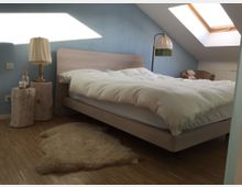 Schlafzimmer unter einer Dachschräge mit hellblauen Wänden, einem Holzbett mit weißer Bettwäsche, einem Schaffell auf einem Holzboden und einem Dachfenster, das Tageslicht hereinlässt. Neben dem Bett stehen Lampen auf Baumstämmen, im Hintergrund ein gepolsterter Sessel.