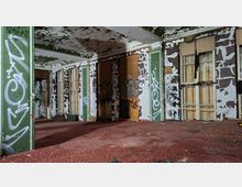 Innenraum eines verlassenen Gebäudes mit abblätternder Farbe an Decke und Wänden, grün-weiß gestrichenen Säulen und mehreren hohen Fenstern, teils mit Brettern vernagelt. Der Raum hat roten Teppichboden und ist mit Graffiti und beschädigten Wandverkleidungen übersät.