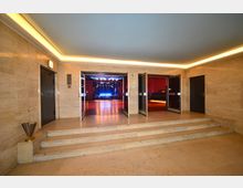 Eingangsbereich zu einem Konzert- oder Veranstaltungssaal mit beige gefliesten Wänden und Treppen, die zu geöffneten Doppeltüren führen. Im Hintergrund ist eine Bühne mit blauer Beleuchtung erkennbar.