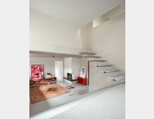 Minimalist split-level living room with white walls, a floating staircase leading to an upper opening, and a white brick corner fireplace. The lower seating area includes modern chairs, a tan rug, low shelving with framed artwork, and a small red cabinet.