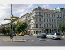 Straßenszene in einer Stadt mit einem historischen, weiß verputzten Gebäude mit Kuppeldach und großen Fenstern an einer Kreuzung. Verkehrsschilder zeigen Richtungen nach Neukölln, Charlottenburg und Schöneberg, während Autos und Fußgänger unterwegs sind.