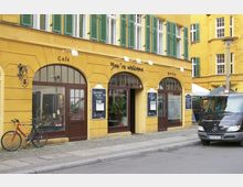 Gelbes Gebäude mit grünen Fensterläden, das ein Café und Bistro beherbergt. Vorne sind braune Türen mit Glas, schwarze Tafeln mit Menüs, ein rotes Fahrrad, und ein dunkler Lieferwagen geparkt.