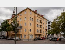 Ecke eines Wohnblocks in einer städtischen Straße mit einem gelb verputzten Mehrfamilienhaus im klassischen Stil, symmetrischen Fenstern und Balkonen. Im Vordergrund stehen Straßenschilder, parkende Autos und Bäume; der Himmel ist wolkig.