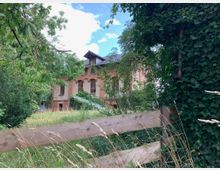 Backsteingebäude mit Satteldach, teilweise von vegetation bedeckt, umgeben von dichtem Grün und einem verwilderten Garten; im Vordergrund ein Holzzaun.
