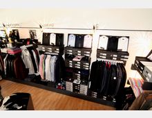 Ein kleiner Laden mit schicker Herrenbekleidung, einschließlich Hemden in schwarzen Schachteln auf Regalen, ordentlich aufgehängten Jacken und Hemden sowie gestapelten Kleidungsstücken auf einem Holzboden. Die Beleuchtung erzeugt dekorative Muster an der weißen Wand.