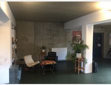 Ein schlichter Innenraum mit Betonwänden und grüner Bodenbeschichtung. Im Raum stehen ein weißer und ein schwarzer Sessel, ein kleiner Tisch mit gemustertem Design, eine große Topfpflanze auf einem Holzständer sowie ein Heizkörper an der Wand. Links ist ein Regal sichtbar, im Hintergrund eine Tür und Fahrradteile.