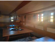 Gemütlicher Innenraum einer rustikalen Stube mit Holzbalkendecke, Holztischen und Sitzbänken. Sonnenlicht fällt durch kleine Fenster mit Holzrahmen, und rustikale Dekoration wie Kerzenleuchter und Fellauflagen schmücken die Tische.