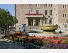 Ein historisches Gebäude mit gelbem Sandstein, großen Fenstern und Blumenkästen mit roten Blumen. Im Vordergrund ein Brunnen mit einer Figur, umgeben von farbenprächtigen Blumenbeeten und einer abgesperrten Grünfläche; links daneben ein bemalter Bärenfigur und Fahrräder.