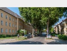 Straßenszene mit mehrstöckigen Wohngebäuden in beige und braun, gesäumt von hohen Bäumen, die Schatten auf die Fahrbahn werfen. Am Straßenrand sind parkende Autos und Gehwege erkennbar, und ein Straßenschild weist auf die Margaretenstraße hin.