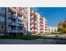 Wohnblock mit fünf Stockwerken in schlichtem Baustil, versehen mit roten Balkonen und teils verputzter Fassade. Im Vordergrund befindet sich ein kleiner Spielplatz mit Sandfläche sowie ein asphaltierter Gehweg, umgeben von Herbstbäumen und Grünflächen.