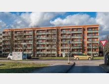 Wohngebäude mit mehreren Etagen in oranger und grauer Farbgestaltung, mit Balkonen entlang der Fassade. Im Vordergrund befinden sich eine Straße, ein Verkehrszeichen und einige geparkte Autos, daneben eine kleine Grünfläche mit kahlen Bäumen.