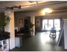 Großzügiger, heller Raum mit industriellem Design und Betonwänden, eingerichtet als Büro oder Atelier. Zu sehen sind Schreibtische mit Bürostühlen vor einer großen Fensterfront, Pflanzen, ein Drucker, Regale und an der Wand Kunstposter.