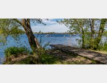 Seeufer mit hölzernem Steg, von Bäumen und Schilf umgeben, im Vordergrund. Im Hintergrund erstreckt sich ein See mit Booten und Menschen auf Stand-up-Paddle-Boards, unter einem klaren blauen Himmel.
