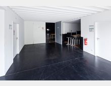 Ein schlichter Innenraum mit weißen Wänden und schwarzem Boden, mehreren Türen sowie einer offenen Abstellfläche mit gestapelten Tischen und Stühlen. Ein Feuerlöscher hängt an der Wand, und eine geöffnete Tür führt in einen dunkleren Raum.