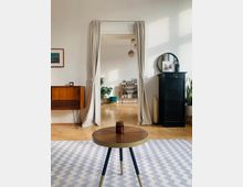 Helles Wohnzimmer mit Holzboden, minimalistisch eingerichtet. Im Vordergrund ein kleiner runder Holztisch auf einem gemusterten Teppich, dahinter eine offene Tür mit weißen Vorhängen, die zu einem weiteren Raum mit Regalen und Pflanzen führt.