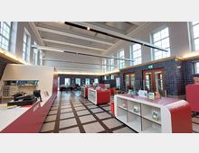 Innenraum einer modernen Bibliothek mit hoher Decke, großen weißen Fenstern und einer Kombination aus roten und weißen Möbeln. Links befindet sich ein Ausleihschalter, in der Mitte Regale mit Prospekten, und im Hintergrund sind Sitzbereiche und Holztüren erkennbar.